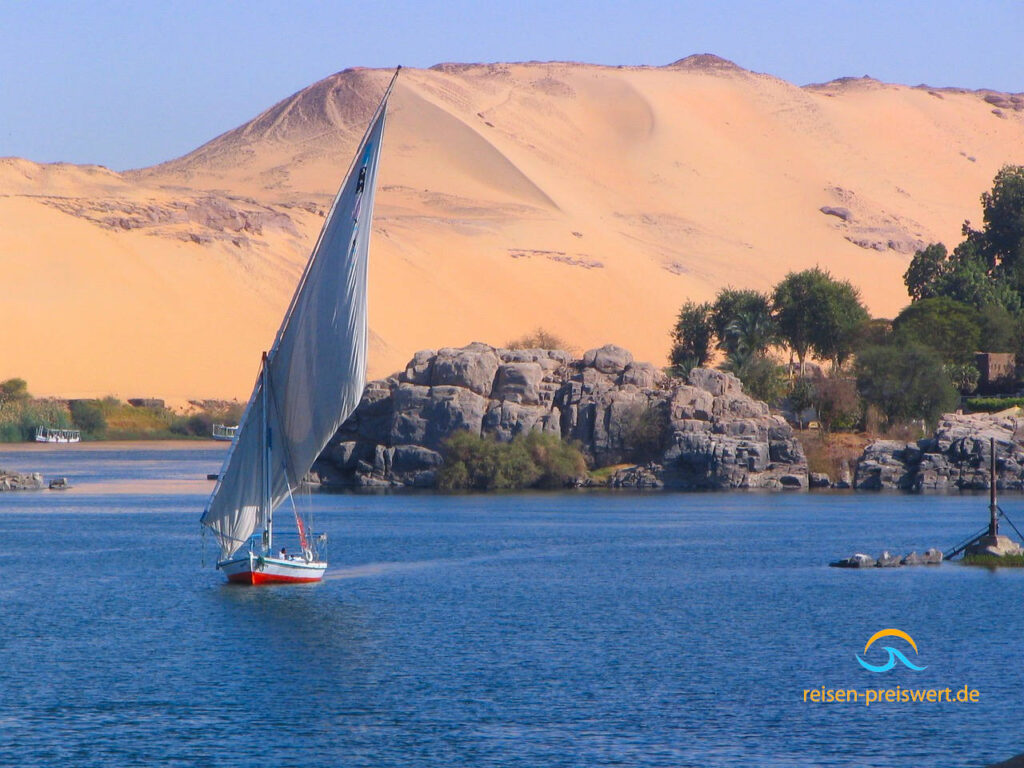 Die Insel Elephantine im Nil - Ägypten. Vorne Wasser mit einem kleinen Segelschiff. Dahinter eine steinige Insel mit Bäumen. Im Hintergrund ein Wüstenberg