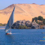 Die Insel Elephantine im Nil - Ägypten. Vorne Wasser mit einem kleinen Segelschiff. Dahinter eine steinige Insel mit Bäumen. Im Hintergrund ein Wüstenberg