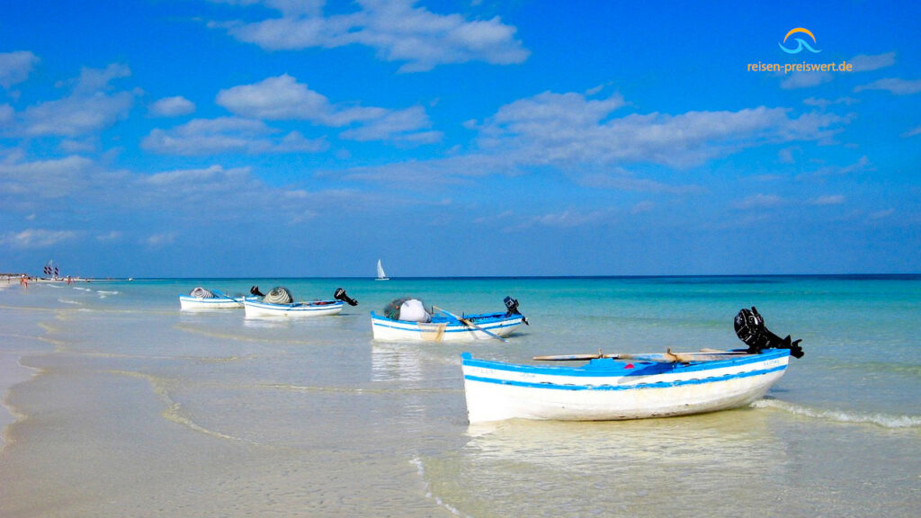 Tunesien: Vier Boote am feinen Sandstrand. Das Wasser ist klar. Der Himmel blau. Die Boote sind blau weiß