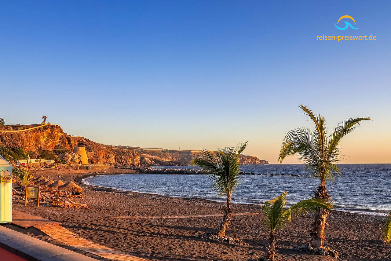 Sonnenuntergang an einem Strand (Bucht) auf Teneriffa. Vorne rechts befinden sch zwei Palmen. Der Himmel ist blau. Am Strand stehen Liegen mit Strohdach.