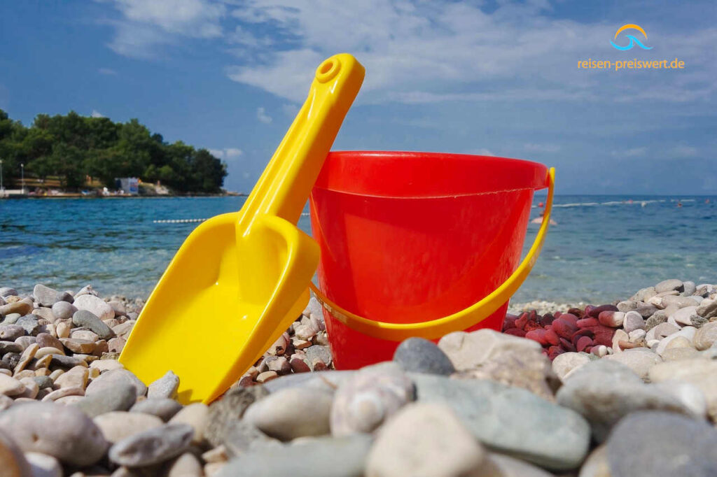 Kinderspielzeug am Meer. Ein roter Platikeimer und eine gelbe Plastikschaufel am Kiesstrand. Das Meer im Hintergrund ist klar, der Himmer leicht bewölkt.