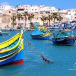 Blick vom Meer in den Hafen einer Stadt auf Malta. Im Vordergrund sind mehrere Fischerboote vor Anker. Im Hintergrund ist eine Promenade mit Gebäuden und einigen Palmen.