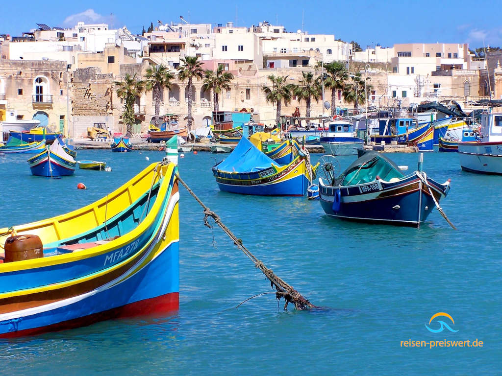 Blick vom Meer in den Hafen einer Stadt auf Malta. Im Vordergrund sind mehrere Fischerboote vor Anker. Im Hintergrund ist eine Promenade mit Gebäuden und einigen Palmen.