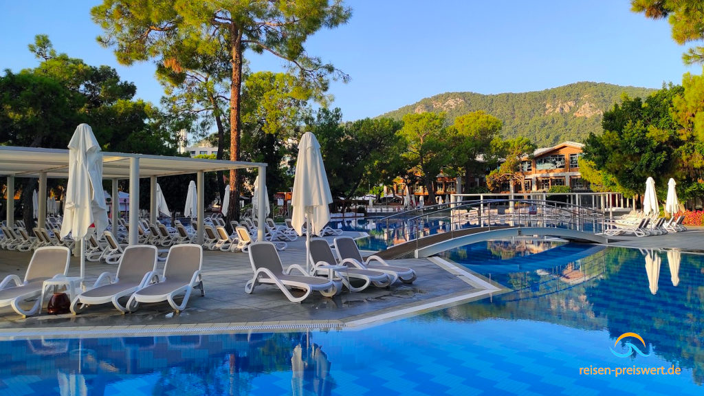 Der Pool im Hotel Rixos Sungate in Kemer. Auf der anderen Seite stehen Liegen und Sonnenschirme. Eine Brücke geht über den Pool und im Hintergrund ist ein Berg mit Bäumen zu erkennen