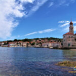 Die Küste vor Sutivan auf Brac in Kroatien - klares Wasser, blauer Himmel. Am Ufer stehen landestypische Gebäude sowie eine Kirche.