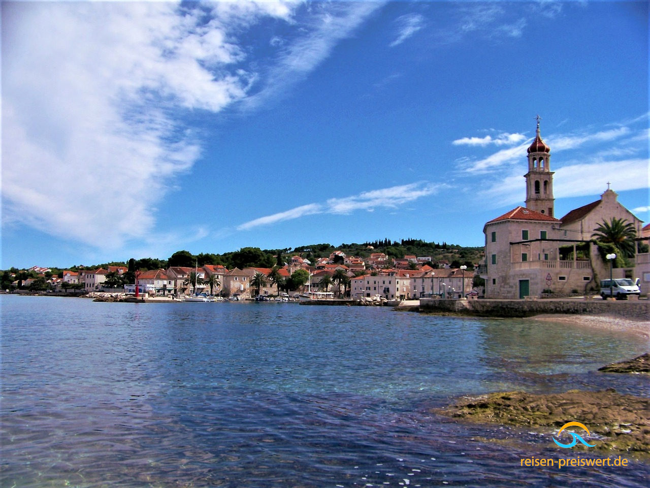 Die Küste vor Sutivan auf Brac in Kroatien - klares Wasser, blauer Himmel. Am Ufer stehen landestypische Gebäude sowie eine Kirche.