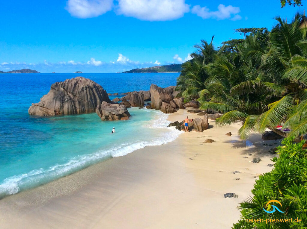 Blick von einer Anhebe auf den feinsandigen hellen Sandstrand. Wellen rollen auf den Strand. Das Meer und der Himmel sind strahlend blau. Im Meer befinden sich Felsen, die zum Klettern einladen