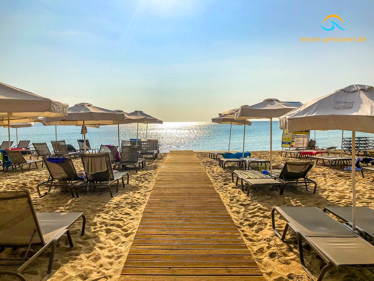 Strand in Bulgarien am Abend. Der Blick geht auf das Meer mit untergehender Sonne. Ein Holzweg führt durch den Sand zum Meer. Links und rechts stehen Sonnenliegen und Sonnenschirme.