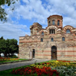 Blick auf die Kirche Christi Pantokrator in Nessebar - Bulgarien. Die Kirche wirkt wie eine kleine Burg mit Rundungen und urigen Fenstern. Im Vordergrund befinden sich bunt blühende Blumenbeete.