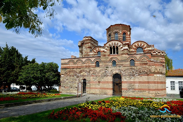 Blick auf die Kirche Christi Pantokrator in Nessebar - Bulgarien. Die Kirche wirkt wie eine kleine Burg mit Rundungen und urigen Fenstern. Im Vordergrund befinden sich bunt blühende Blumenbeete.